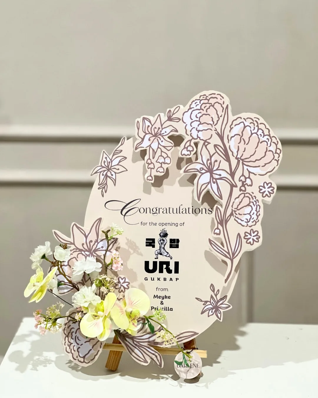 Contoh papan krem bertulisan Congratulations dengan logo penerima, nama pengirim, dan bunga putih-kuning